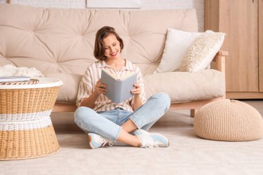 Genç, güzel bir kadın kitap okuyor ve oturma odasındaki kanepenin yanında oturuyor.