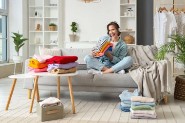 Kulaklıklı genç bir kadın farklı giysiler katlıyor ve evdeki koltukta oturuyor.