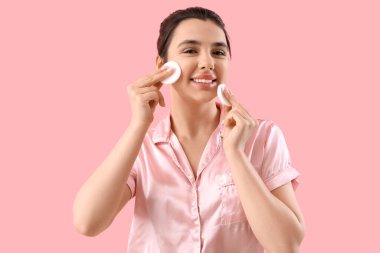 Genç bir kadın pembe arka planda pamuk pedlerle makyaj yapıyor.