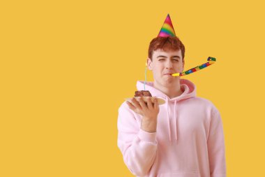 Parti üfleyicisi ve sarı arka planda doğum günü pastası olan genç bir adam.