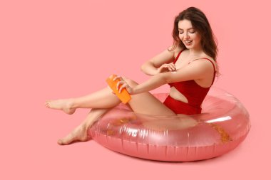 Yüzme yüzüklü genç bir kadın pembe arka plana güneş kremi sürüyor.