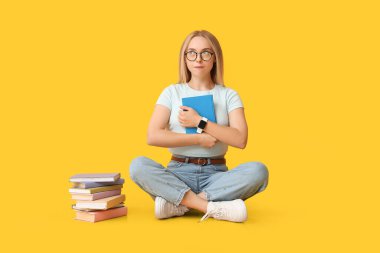 Sarı arka planda oturan bir sürü kitabı olan güzel genç bir kadın.