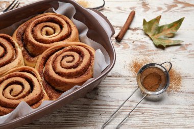 Beyaz ahşap zemin üzerinde lezzetli tarçın ruloları ve sonbahar yaprağı pişirme tabağı