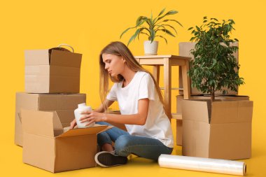 Güzel genç bayan, sarı arka plandaki karton kutularda eşyalarını topluyor. Taşınma konsepti