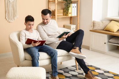Baba ve küçük oğlu evdeki kanepede kitap okuyorlar.