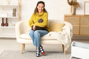 Evde elektrikli kaloriferin yanında tatlı kedisi olan mutlu olgun bir kadın oturuyor.