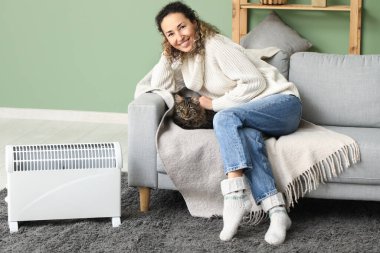 Evde elektrikli kaloriferin yanında tatlı kedisi olan mutlu olgun bir kadın oturuyor.