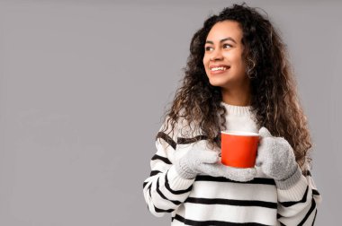 Gri arka planda sıcak sıcak bir bardak şarapla sıcak eldivenler giymiş mutlu Afro-Amerikalı kadın.