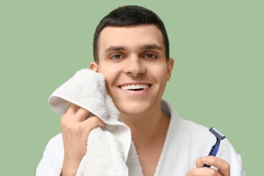 Tıraş bıçağı ve yeşil arka planda havlusu olan bornozlu yakışıklı bir gencin portresi.