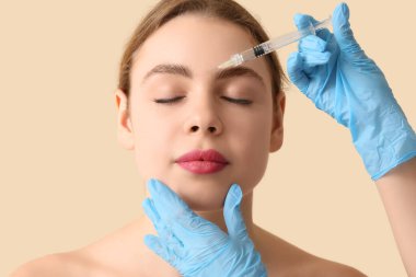 Güzel genç bir kadın bej arka planda yüzüne enjeksiyon yaptırıyor.