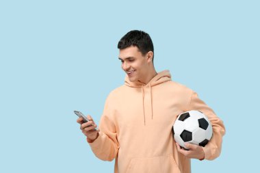 Mavi arka planda futbol topu ve cep telefonu olan genç bir adam.
