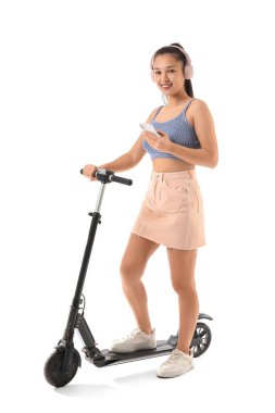 Kulaklıklı, modern elektrikli scooter 'ı ve beyaz arka planda cep telefonu olan güzel bir Asyalı kadın.
