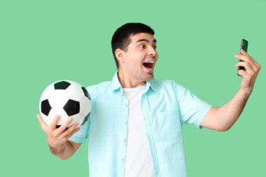 Yeşil arka planda futbol topu ve cep telefonu olan mutlu genç bir adam. Spor bahsi konsepti