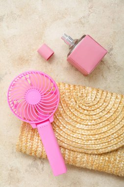Portatif pembe elektrikli vantilatör, hasır çanta, parfüm ve gri grunge arka planda palmiye yaprakları.