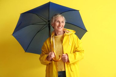 Sarı arka planda şemsiyesi olan yağmurluk giymiş güzel olgun bir kadın.
