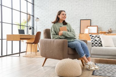 Kitabı olan mutlu genç bir kadın evdeki koltukta oturuyor.