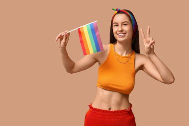 Göbeğinde piercing ve LGBT bayrağı olan mutlu genç bir kadın bej arka planda zafer jesti gösteriyor.