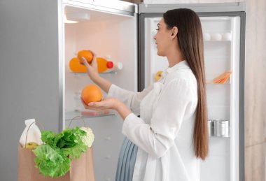 Genç hamile kadın mutfaktaki buzdolabına portakal koyuyor.