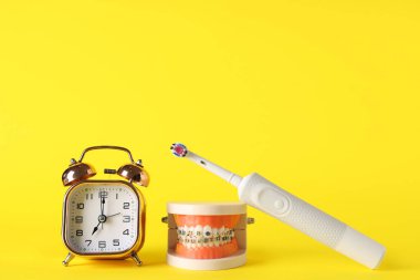Alarm saati, diş telli çene modeli ve sarı arkaplanda elektrikli diş fırçası
