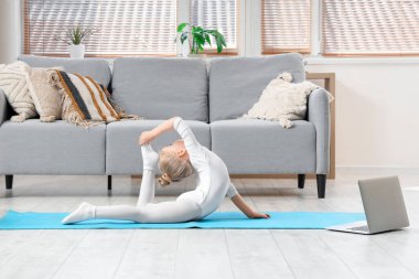 Cute little girl doing gymnastics while watching online tutorial video on laptop at home