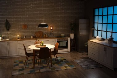 Parlayan lambaları, beyaz tezgahları, fırını, sandalyeleri ve akşam yemeği masası olan mutfağın içi.