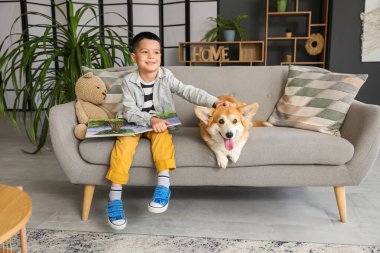 Küçük mutlu Asyalı çocuk evde kanepede tatlı Corgi köpeğiyle kitap okuyor.