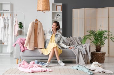 Genç, hayal kırıklığına uğramış bir kadın ceketini tutuyor ve evdeki farklı kıyafetlerin yanında kanepeye oturuyor.