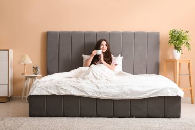 Güzel, genç bir kadın, bir fincan çayla rahat bir yatakta yatıyor.