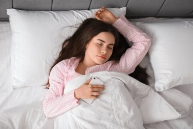 Pretty young woman with smartphone sleeping in cozy bed clipart