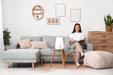 Genç kadın rahat koltukta dergi okuyor modern nemlendiricinin yanında şık oturma odasında.