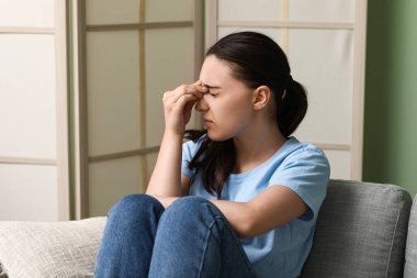 Baş ağrısı çeken ve evdeki kanepede oturan genç bir kadın.