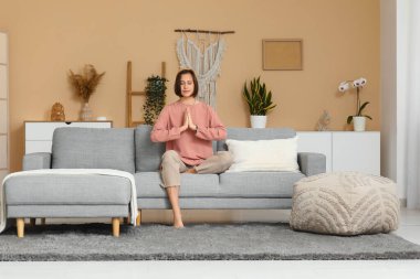 Evdeki kanepede oturmuş meditasyon yapan genç bir kadın.