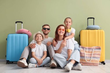 Yeşil duvarın yanında bavullu turist ailesi oturuyor.