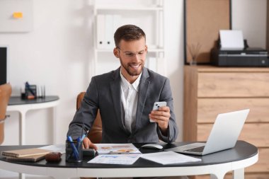 İş yerinde gözlüklü genç işadamı cep telefonu kullanıyor. Ofisinde belgeler ve dizüstü bilgisayar var.