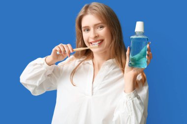 Genç bir kadın dişlerini fırçalıyor ve elinde mavi arka planda bir şişe gargara tutuyor.