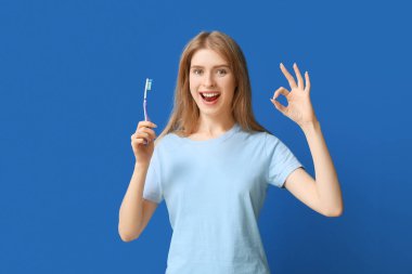 Diş fırçalı genç bir kadın mavi arka planda iyi bir jest gösteriyor.