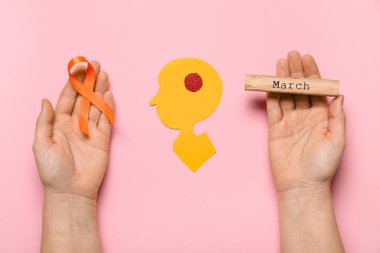 Turuncu kurdeleli kadın eller, marş sözü ve pembe arka planda kağıt insan kafası. Çoklu Skleroz Farkındalık Ayı