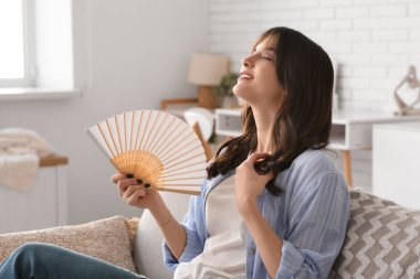 Elinde yelpaze olan genç bir kadın oturma odasında kanepede oturuyor.