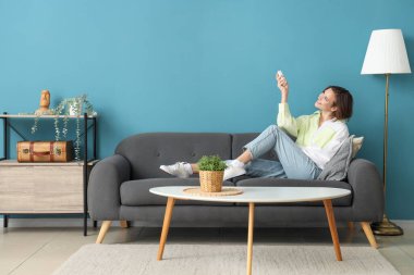 Mavi duvarın yanındaki kanepede duran klimalı uzaktan kumandalı gülümseyen genç kadın.