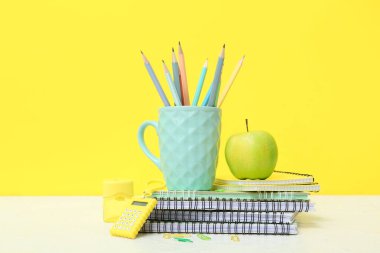 Bardaktaki kalemler, defterler, okul malzemeleri ve yeşil elma sarı arka planda.