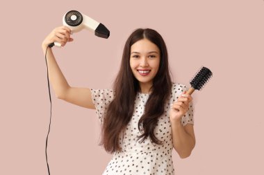 Saç kurutma makinesi ve pembe arka planda fırçası olan güzel bir Asyalı kadın.