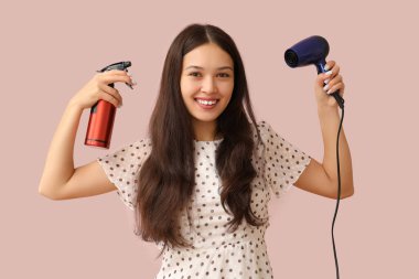 Saç kurutma makinesi ve pembe arka planda spreyi olan güzel bir Asyalı kadın.