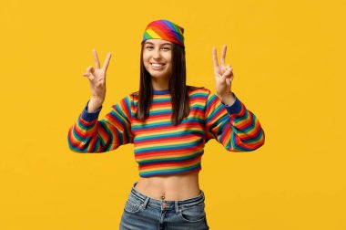 Gökkuşağı süveter giyen, göbeğinde piercing olan genç ve güzel bir kadın sarı arka planda zafer jesti yapıyor.