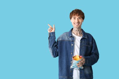 Mavi arka plandaki bir şeyi işaret eden patates cipsli genç dövmeli adam.