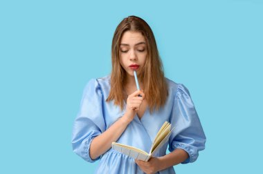 Mavi arka planda not defteri ve kalemi olan düşünceli genç bir kadın. Hatırlatma konsepti