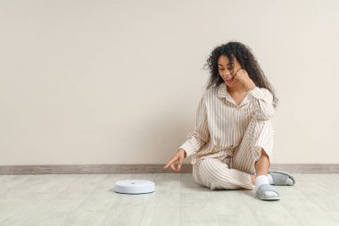 Pijamalı genç Afro-Amerikalı kadın bej duvar kenarındaki robot elektrik süpürgesini işaret ediyor.