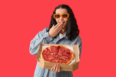 Güneş gözlüklü genç bir kadın elinde kırmızı arka planda lezzetli pizzayla karton bir kutu tutuyor.