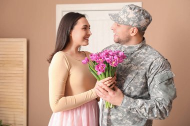 Man in military uniform hugging his wife with flowers at home. Valentine's Day celebration