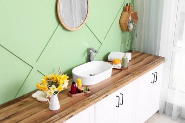 Modern lavabo, banyo malzemeleri ve çiçekler banyodaki yeşil duvarın yanındaki ahşap çekmecede.