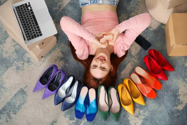 Genç bir kadın dizüstü bilgisayarla, cep telefonuyla yerde yatıyor ve odada topuklu ayakkabılar arasında kalp yapıyor.